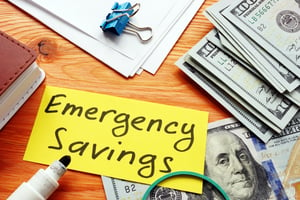 A sign among stacks of money titled Emergency Savings | First Alliance Credit Union