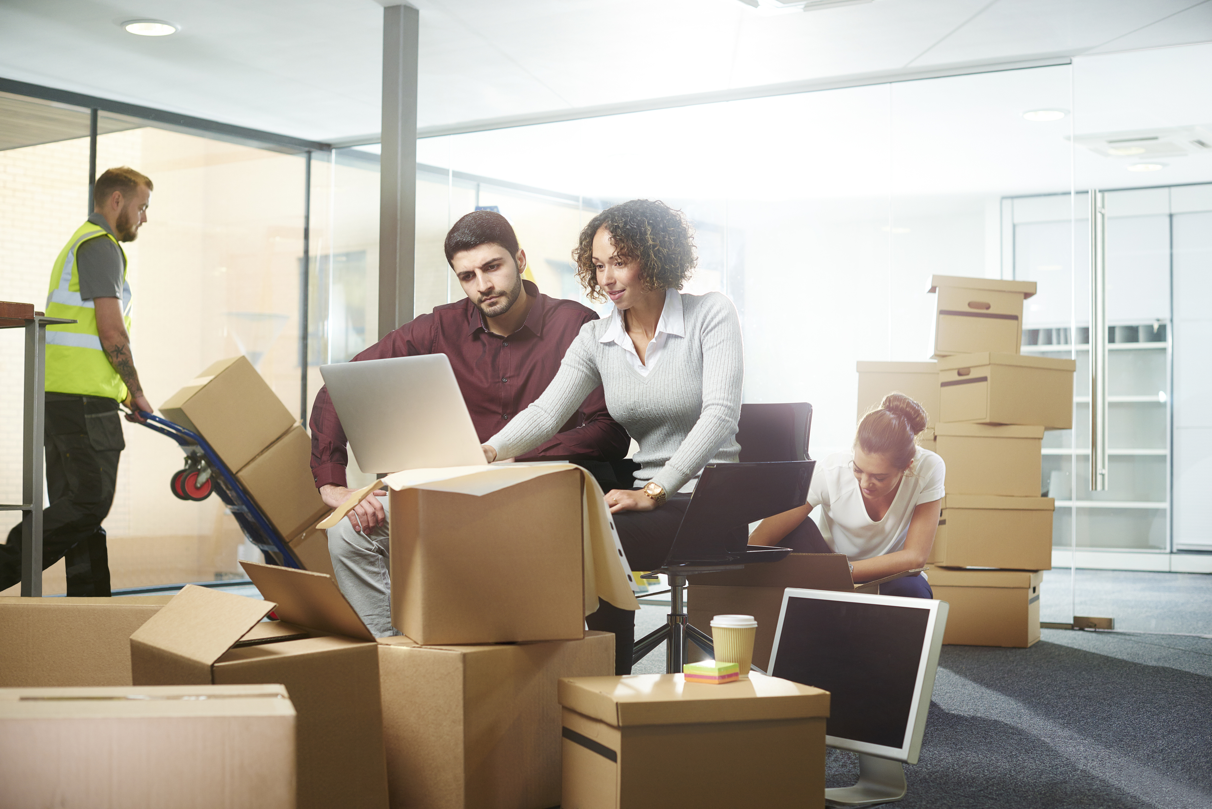 A couple on their laptop in the middle of a bunch of moving boxes as a mover works behind them. 